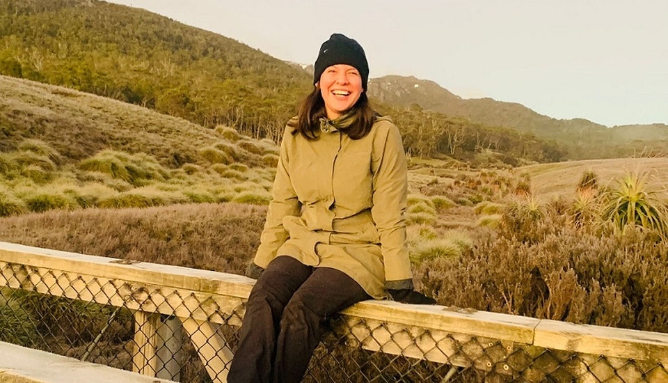 Australian alumna Jess sitting on a fence with a green mountain behind her