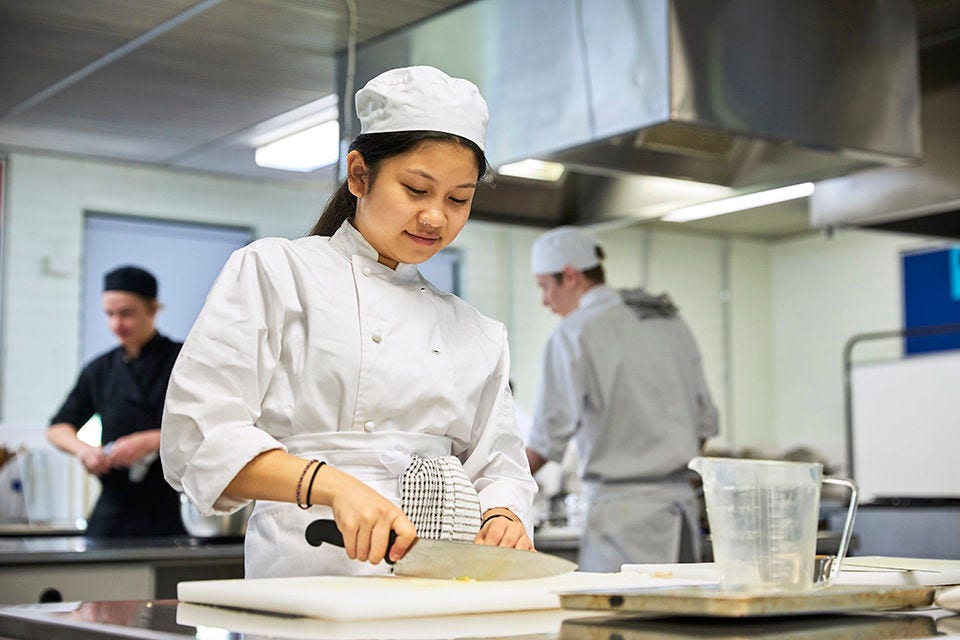 Two students dressed in chef whites cooking in a kitchen.