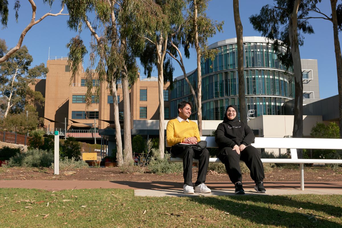 Two international students sitting on a bench with their campus building in the background