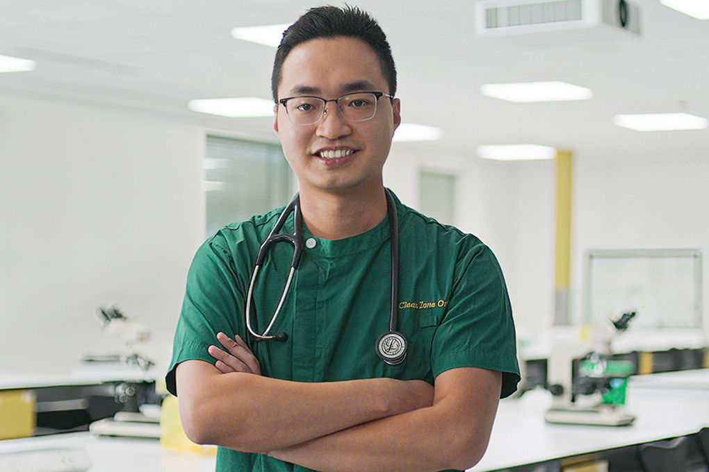 Dr Kwan Leung Chia (Hong Kong) has a passion for integrating Eastern and Western medicine.  He is standing in a lab wearing hospital scrubs and has a stethoscope around his neck. He is smiling at the camera with his arms crossed.