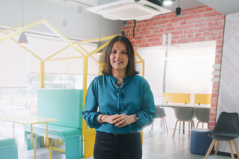 Dr Reema Joshi stands smiling at the camera in an office breakout space. 