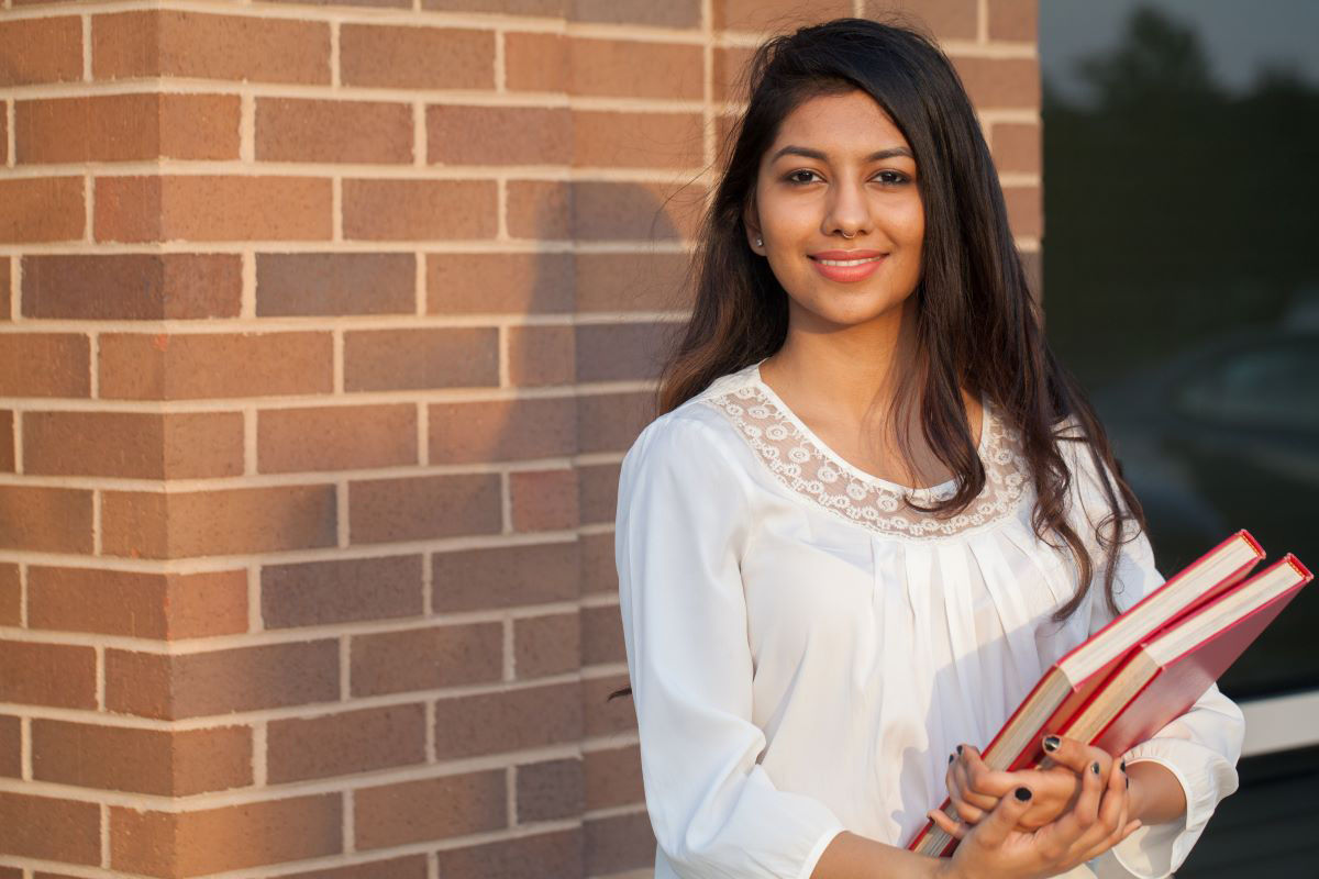 A young female international student is smiling at the camera and holding books