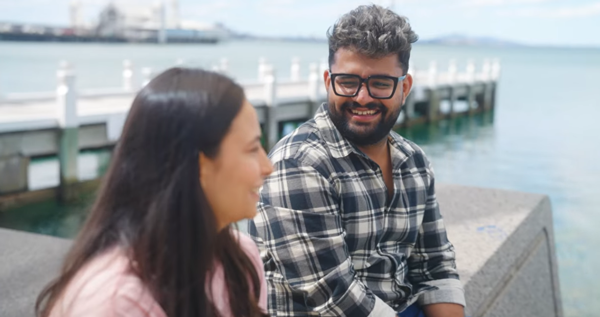 Vish sitting by the water with a pier in the background, chatting to a friend