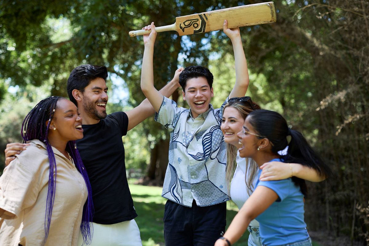 Five students laughing while one holds a cricket bat in the air