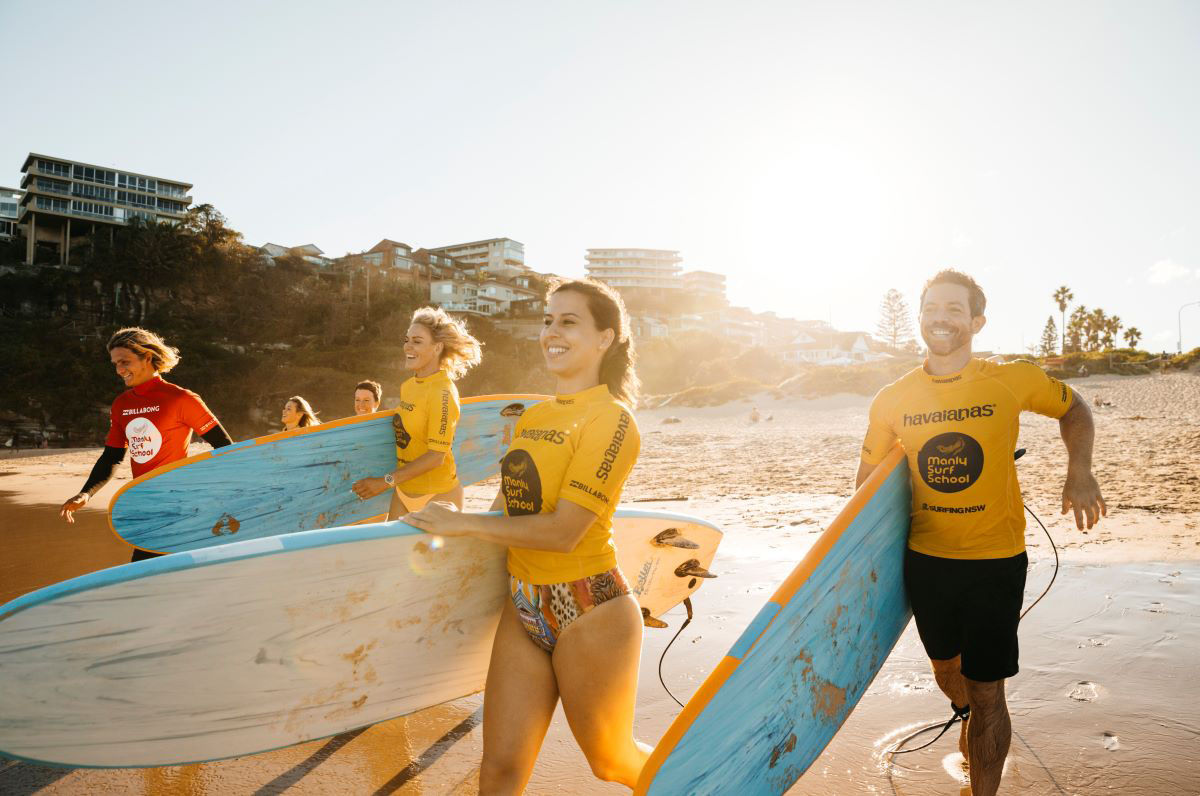 A group of happy students run along a beach carrying surfboards