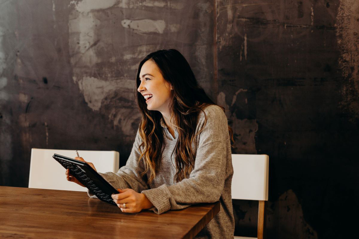 A young woman is sitting at a desk smiling and holding a pen and tablet device 