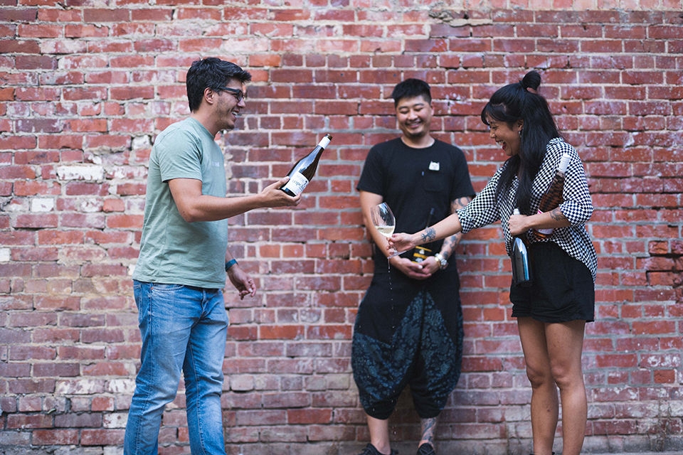 Hyemi, a female international student from South Korea and two fellow male international student alumni - LT (Hu Lui) from China, and Henri from Brazil, are pouring a bottle of wine into glasses..