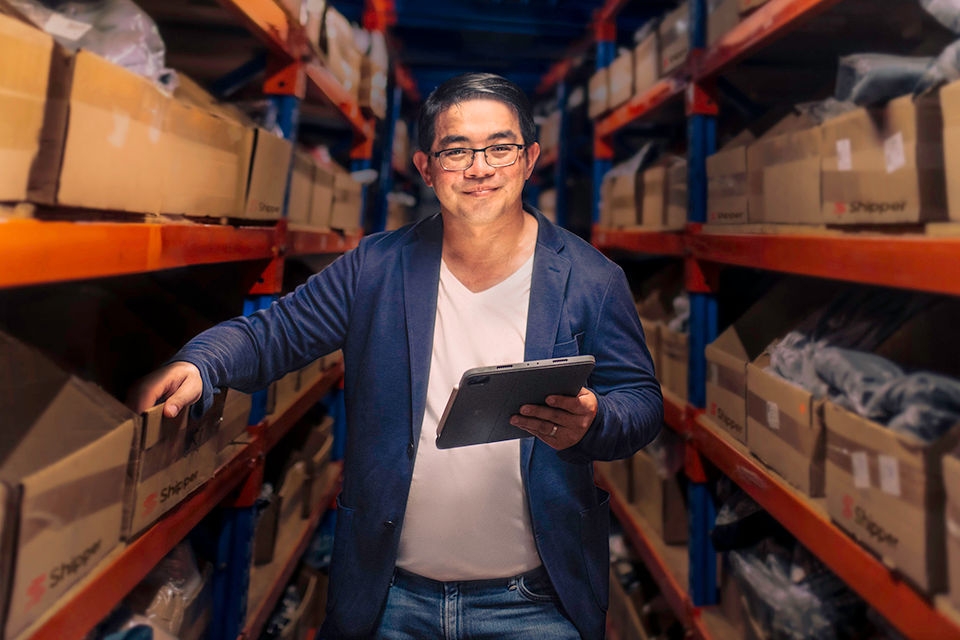A man, Budi Handoko, stands in a Shipper warehouse surrounded by carboard boxes> He is holding a tablet device and smiling. 