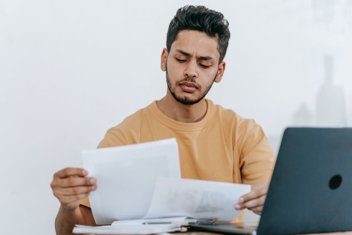 A male international student is looking through paperwork and sitting at his laptop